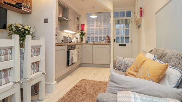 A compact living space with a kitchenette, a sofa with patterned cushions, and a small wooden bench. - Little Gem (Whitby)