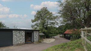 A rustic building with a stone exterior and a metal roof, a gravel driveway, and a fenced area with a small red-roofed structure. - The Snug (Chepstow)