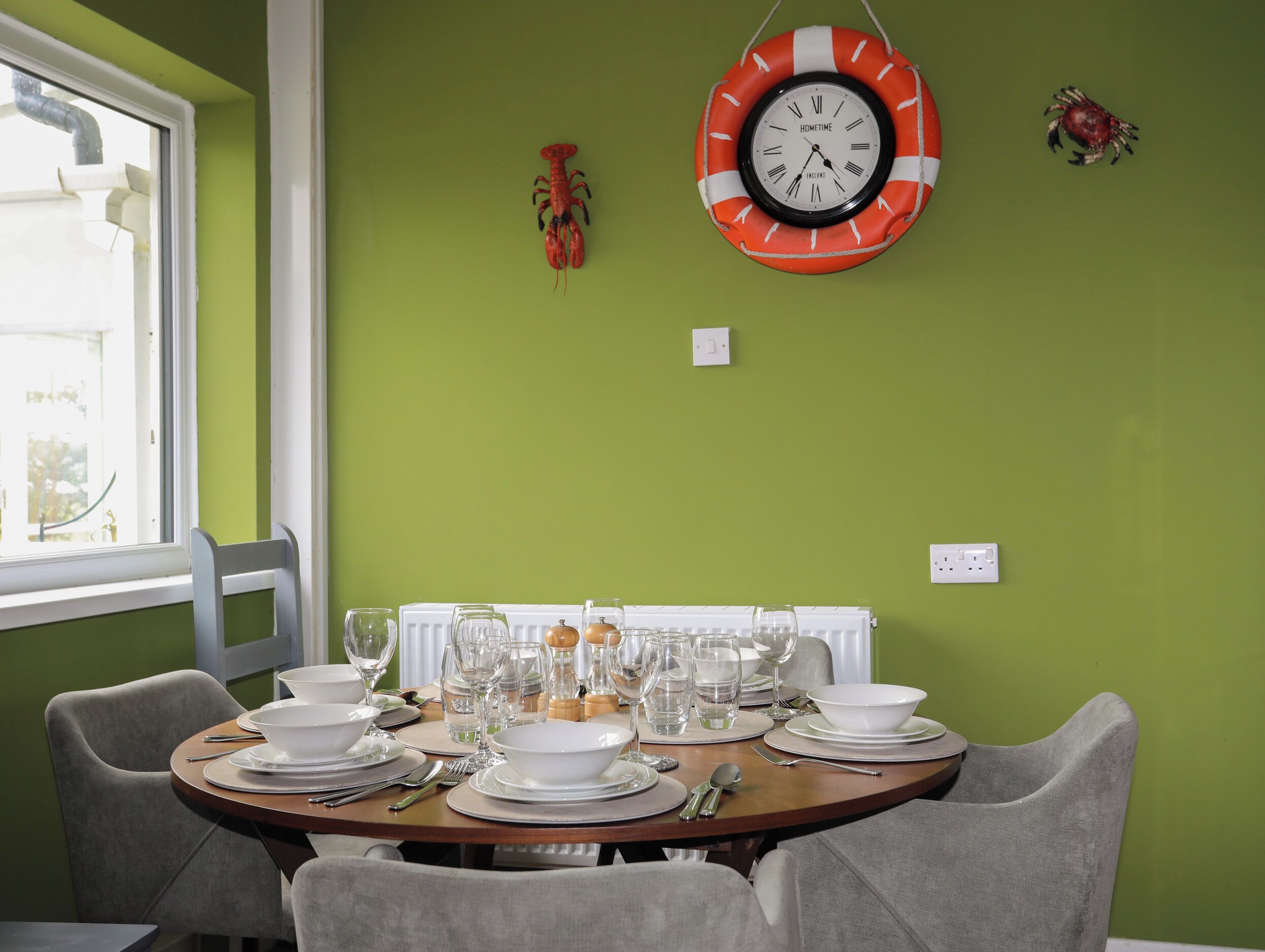 Una mesa de comedor dispuesta con platos blancos, vasos y cubiertos, rodeada de sillas grises, frente a una pared verde con un reloj y una decoración de langosta.