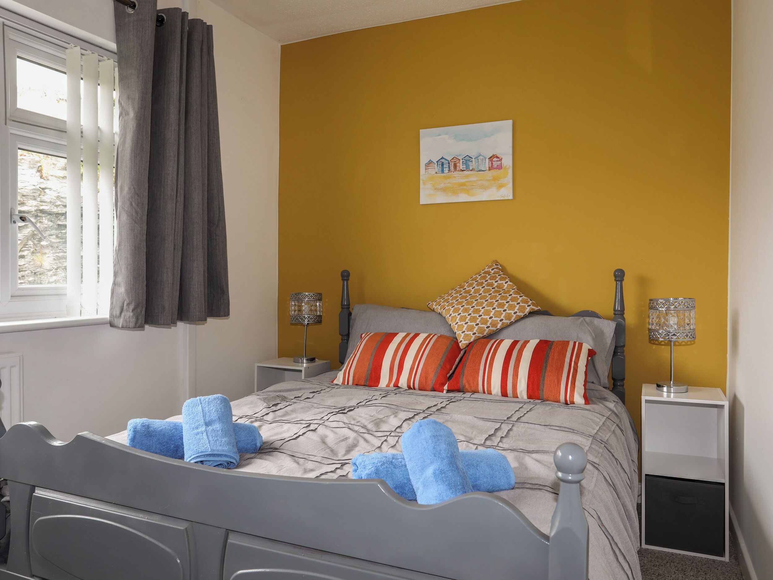 Une chambre à coucher avec un lit, deux tables de chevet, une fenêtre avec des rideaux, un tableau au mur et une petite table de nuit avec une lampe.