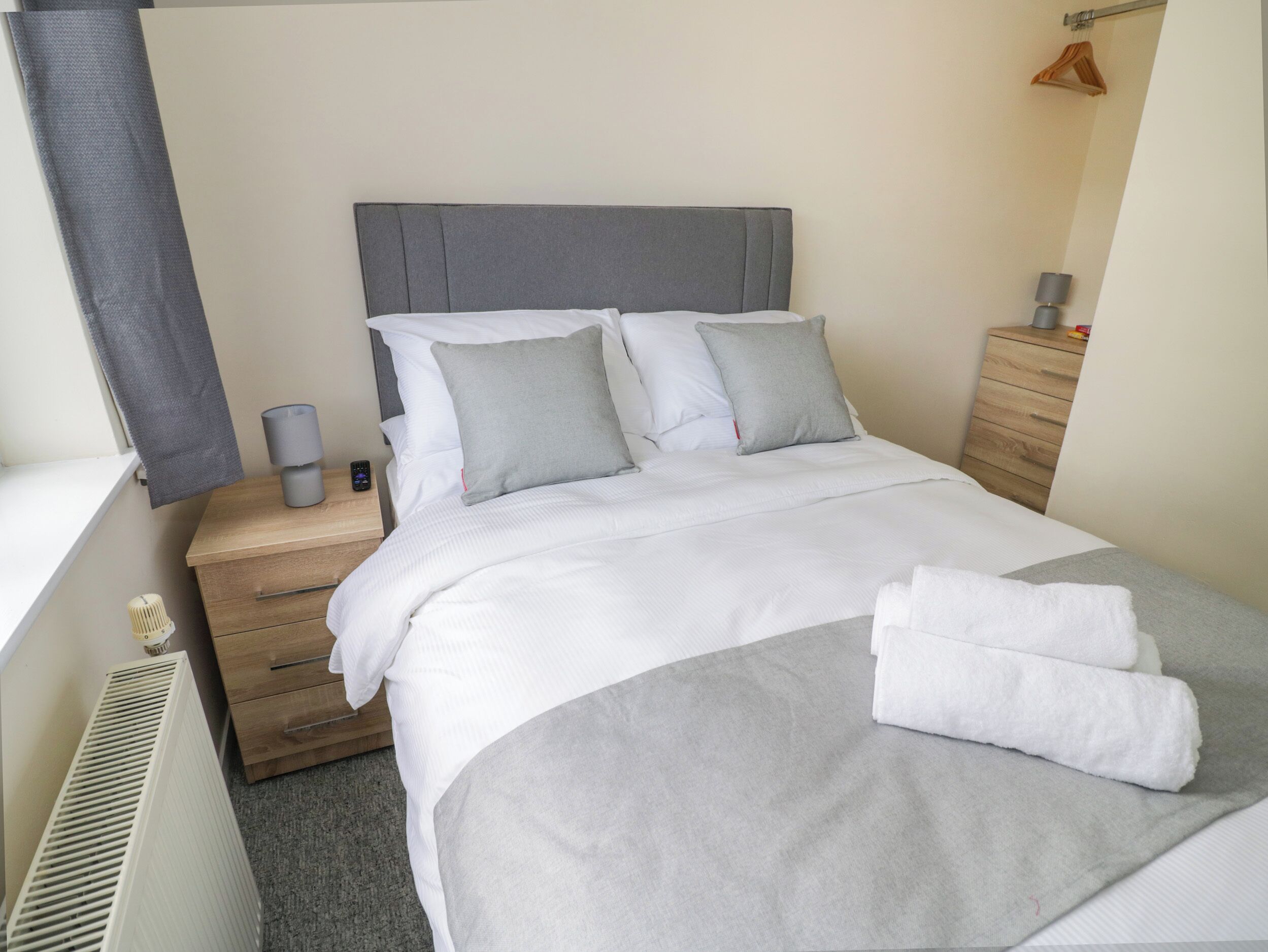 Een netjes opgemaakt bed met wit-grijs beddengoed, een grijs hoofdbord en een houten nachtkastje met een lamp en afstandsbediening.