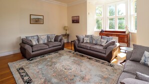 A living room with two sofas, a patterned rug, a piano, and a window with a view of greenery. - North Wing (Belford)