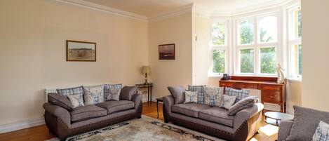 A living room with two sofas, a patterned rug, a piano, and a window with a view of greenery.