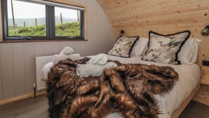 Une chambre confortable avec un plafond en bois, une grande fenêtre donnant sur la verdure, un lit avec du linge de maison blanc et un plaid en fourrure, ainsi que deux coussins ornés d’un motif d’oiseau.