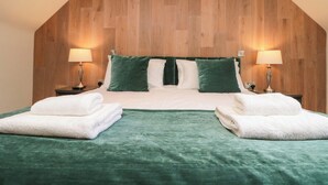 A bedroom with a wooden sloped ceiling, a bed with green and white bedding, two bedside lamps, and two folded white towels on the bed. - Clematis Cottage (Whitby)