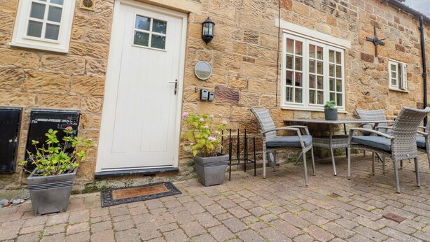 A stone building with a white door, a window, and a doorbell. There are potted plants and outdoor furniture. - Clematis Cottage (Whitby)