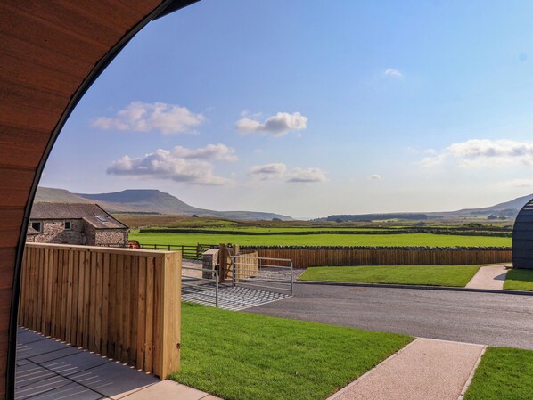 A view through a wooden-framed window of a rural landscape with green fields, a stone building, and a mountain in the distance.