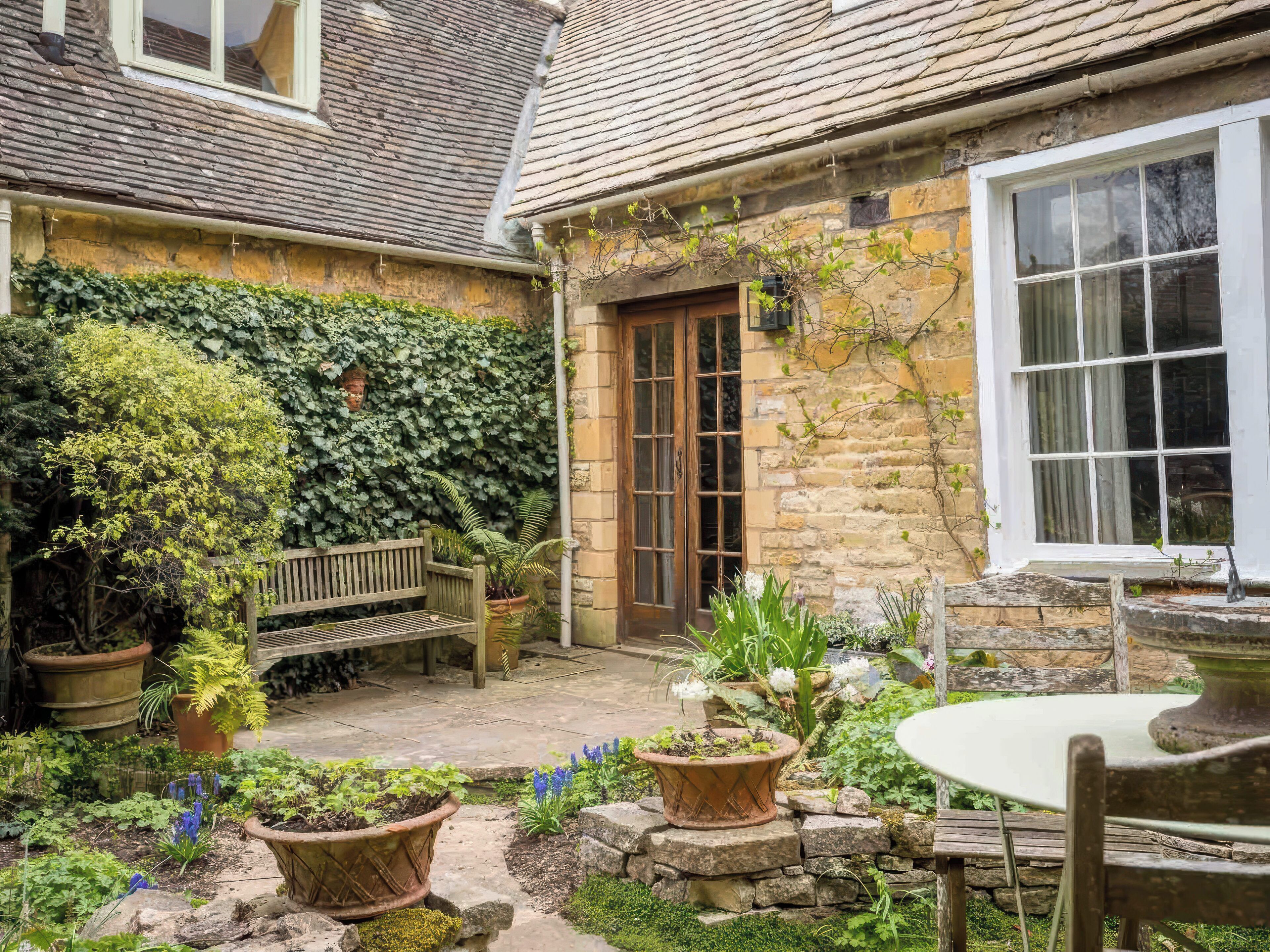 A stone house with a wooden bench, potted plants, and a garden area with a table and chairs.