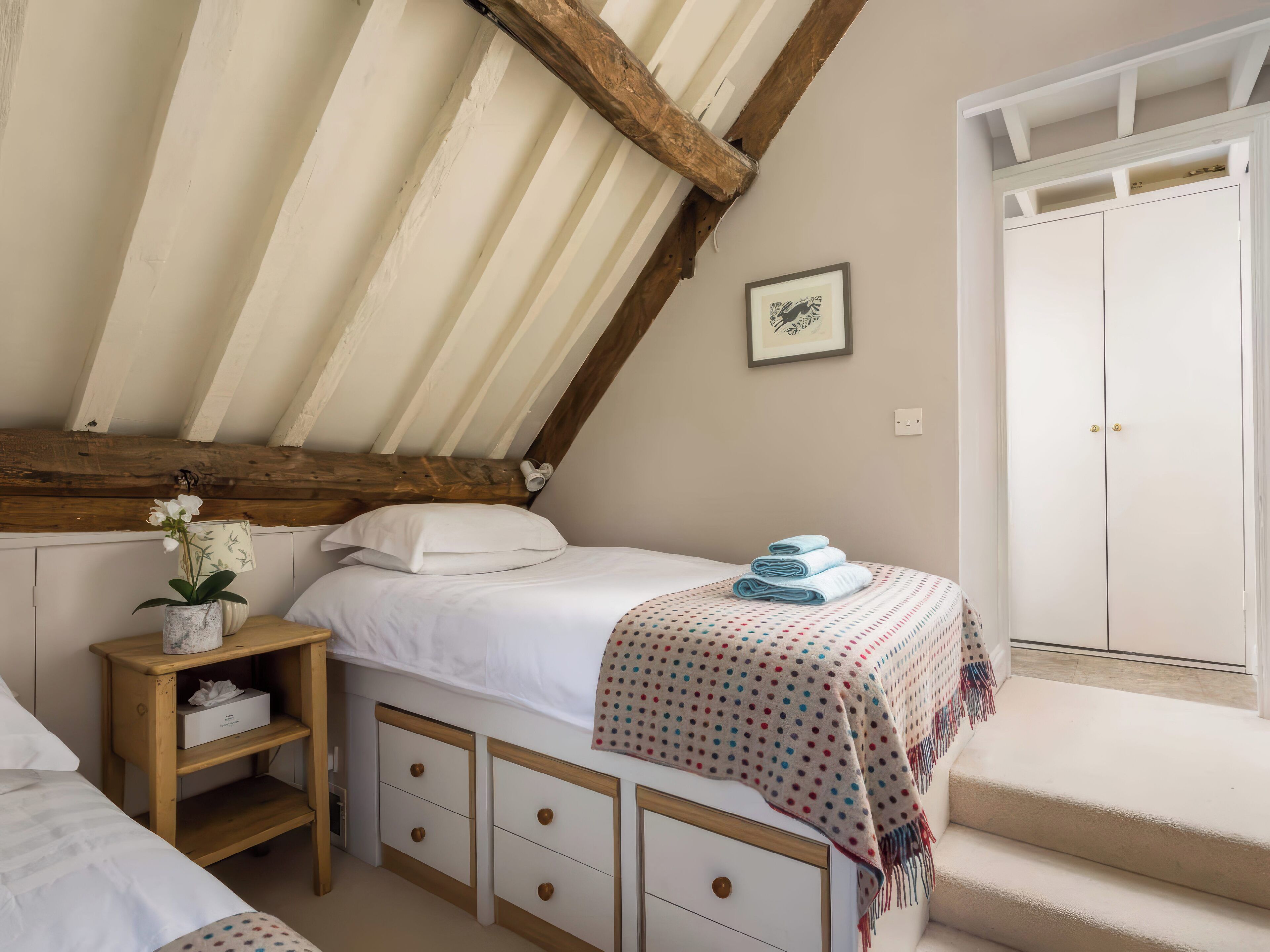 A bedroom with a sloped ceiling, a bed with a polka dot blanket, a bedside table with a vase of flowers, and a wardrobe.