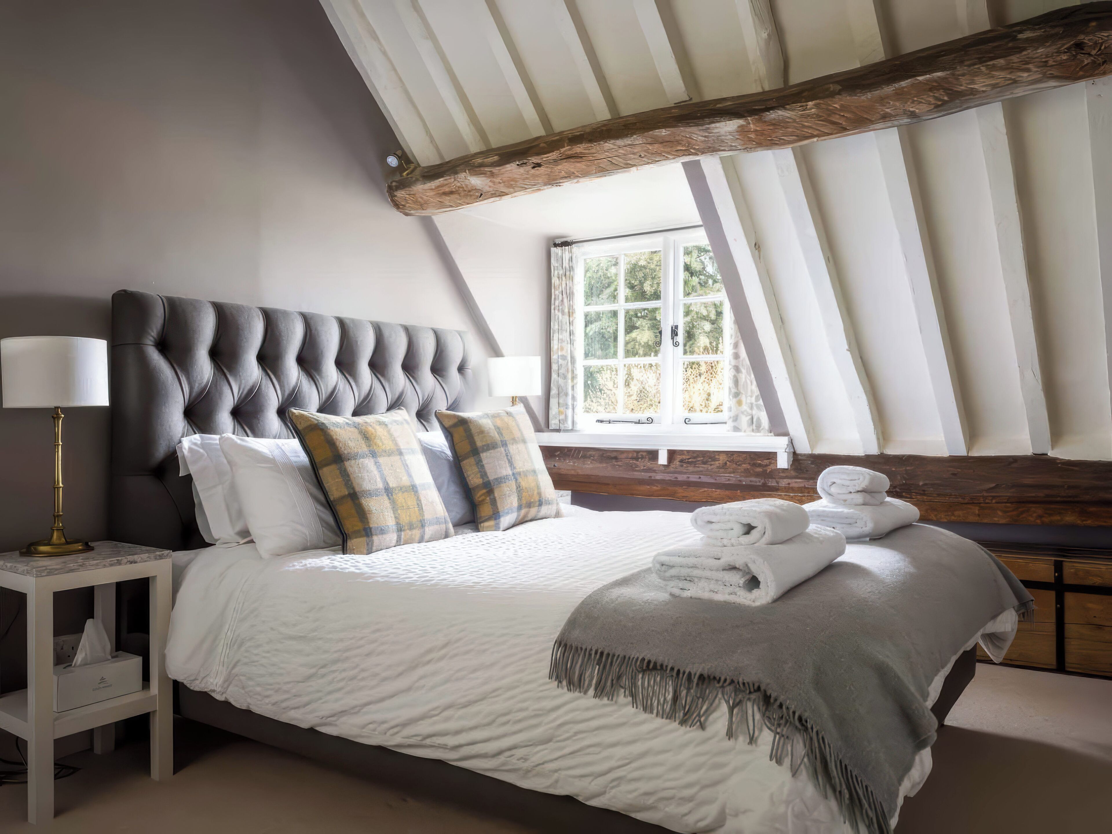 A bedroom with a tufted headboard, a bed with white and gray bedding, a bedside table with a lamp, and a window with a view of greenery.