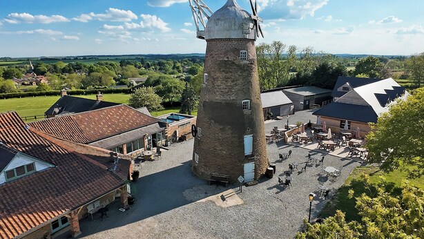 A historic tower with a clock, surrounded by traditional buildings and a courtyard with outdoor seating. - The Oak Room - Windmill Country Retreats (Melton Mowbray)