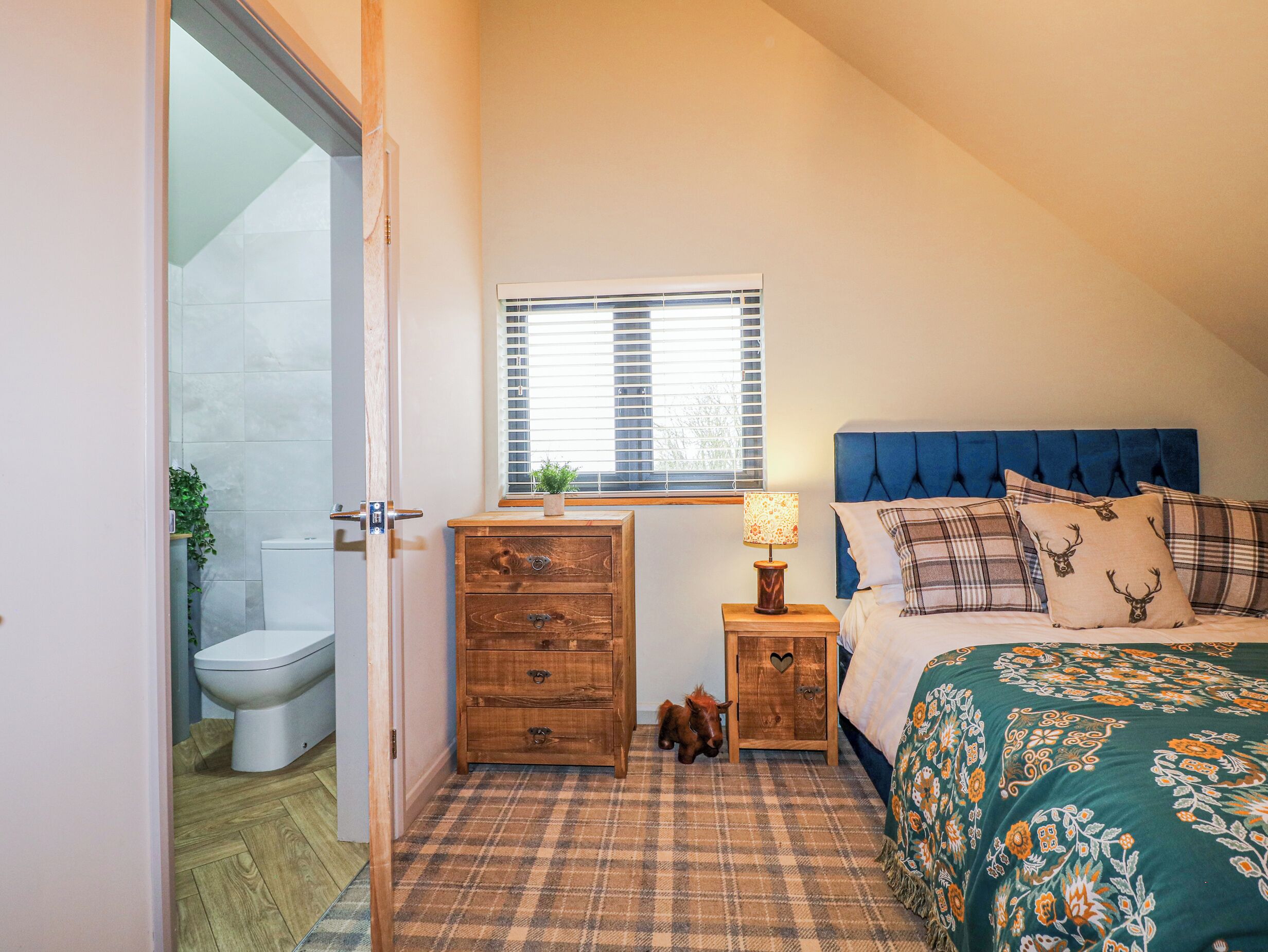 Een slaapkamer met een bed, een houten dressoir, een nachtkastje met een lamp, een raam en een deur naar een badkamer.