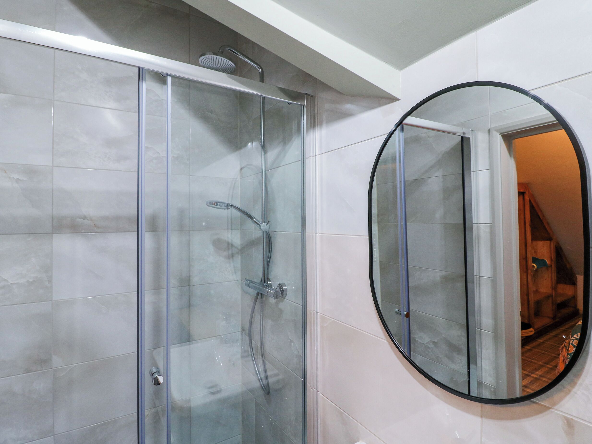 Un baño moderno con cabina de ducha de vidrio, un espejo redondo y un pasillo visible.
