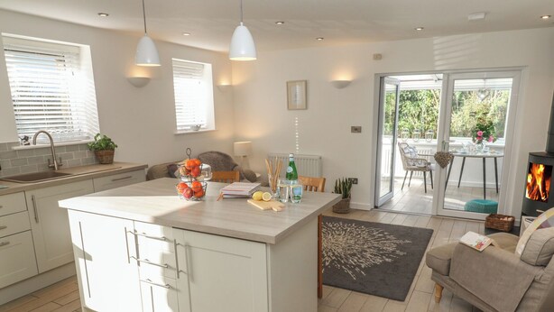 A modern kitchen with a central island, white cabinetry, and a dining area with a sofa and table. - The Duck House (Penzance)