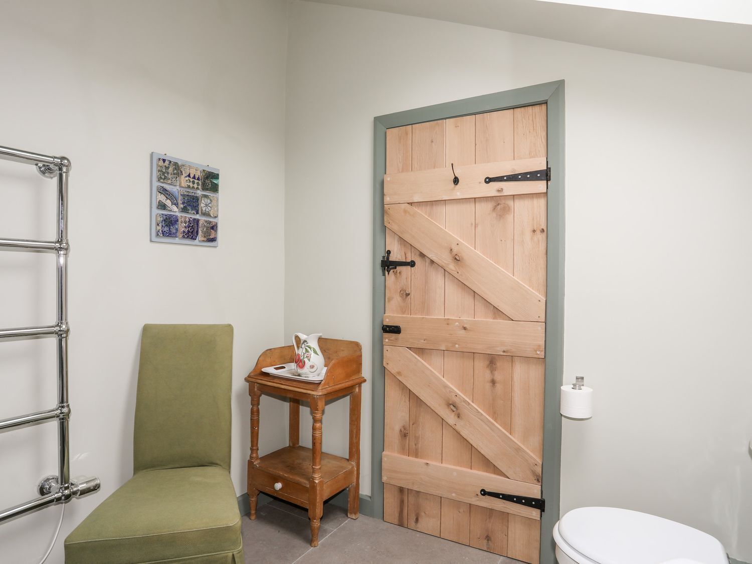 A bathroom with a wooden door, a green chair, a small wooden table with a pitcher, and a metal towel rack.