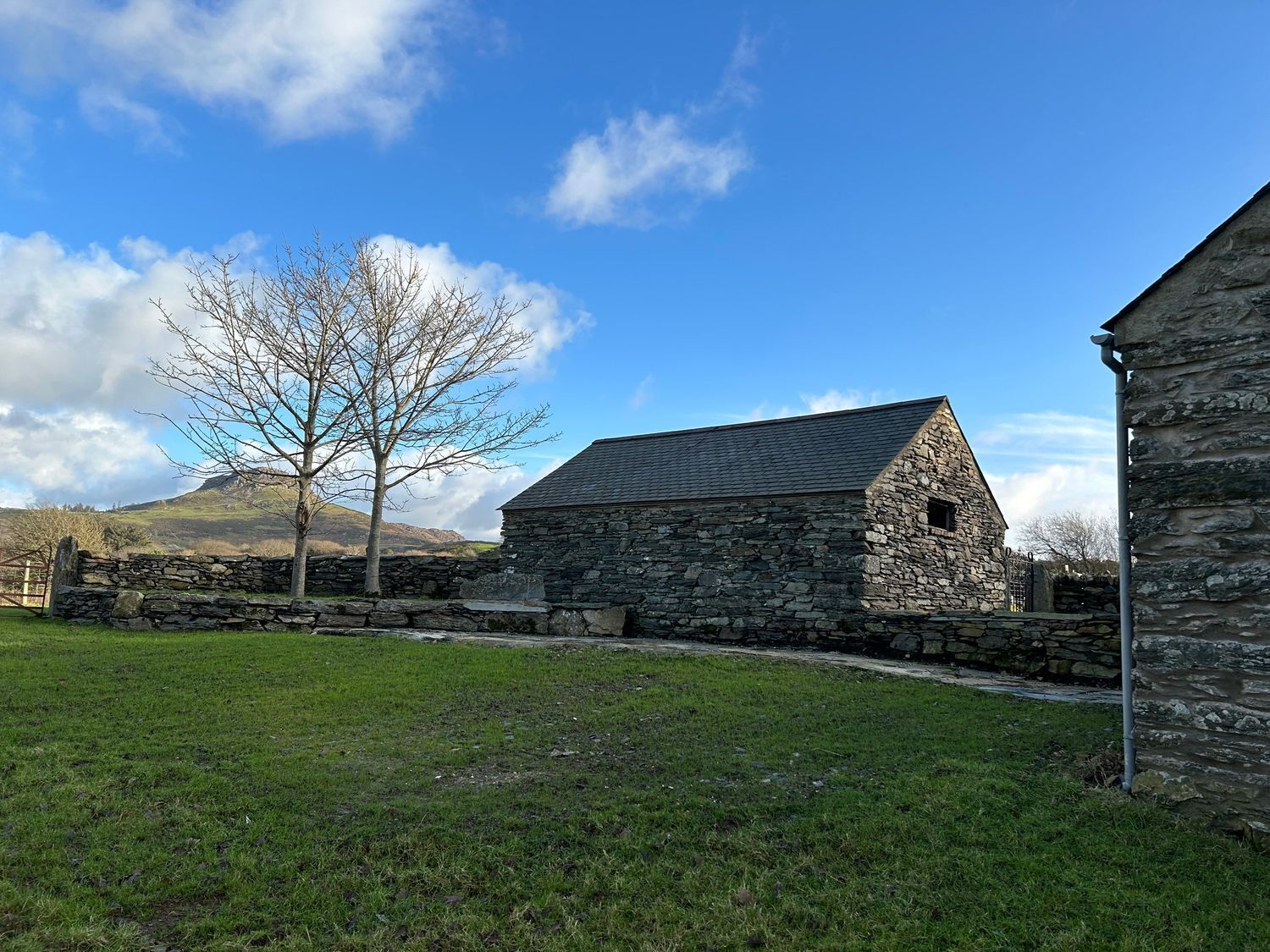 A stone building with a sloped roof, surrounded by a stone wall and a grassy field.