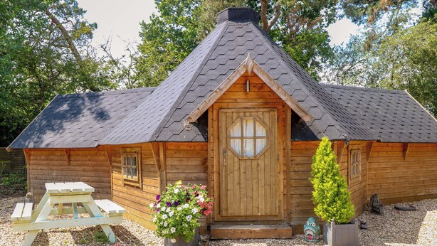 A wooden cabin with a hexagonal window, a picnic table, and potted plants. - Enchanted Cottage (King's Lynn)