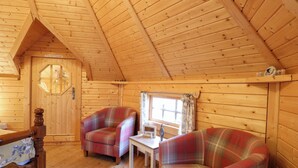 A cozy cabin interior with wooden walls and a sloped ceiling. There are two plaid chairs, a wooden table, and a small window with curtains. - Enchanted Cottage (King's Lynn)