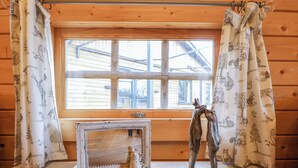A wooden cabin room with a window, curtains with animal prints, a rustic table, and a decorative sculpture. - Enchanted Cottage (King's Lynn)