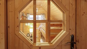 A hexagonal window with a wooden frame, revealing a room with a bunk bed, a lamp, and a clock. - Enchanted Cottage (King's Lynn)