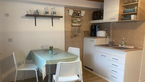 Une cuisine compacte équipée d’un four à micro-ondes, d’une machine à café et d’un petit réfrigérateur. On y trouve une table à manger avec des chaises, et une étagère au-dessus, où sont rangés les ustensiles de cuisine.