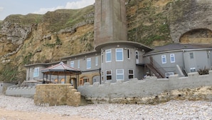 Ein Küstengebäude mit einem markanten Turm, Steinmauern und einem Strand im Hintergrund.