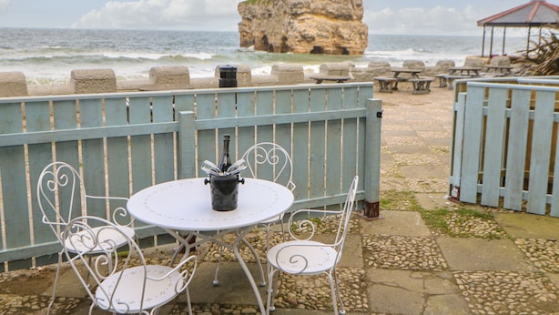 Eine Terrasse am Meer mit Tisch und Stühlen, einer Weinflasche und Blick auf das Meer und eine große Felsformation.
