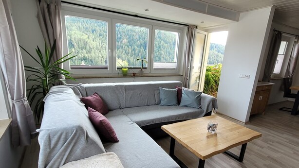 Un salón moderno con un sofá modular gris, una mesa de centro de madera y ventanas amplias que dan a una zona boscosa.