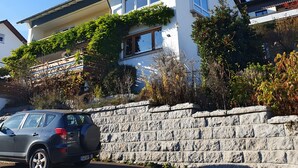 Una casa de dos pisos con un muro de piedra en el frente, un coche estacionado y un cielo azul despejado.