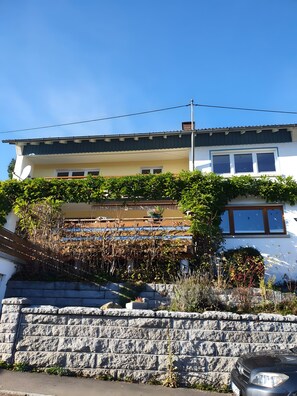 Una casa a due piani con un muro in pietra in primo piano, un balcone con piante e un cielo azzurro limpido.