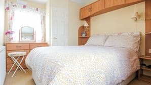 A bedroom with a bed, a dresser, a mirror, a window with curtains, and a nightstand with a lamp. - 120 Caravan (Great Yarmouth)