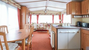 A compact RV kitchen with wooden cabinets, a small refrigerator, and a dining area with a table and chairs. - 120 Caravan (Great Yarmouth)