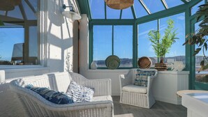 A sunlit room with wicker furniture, a large window, and decorative items. - Swn Y Wylan (Pentraeth)