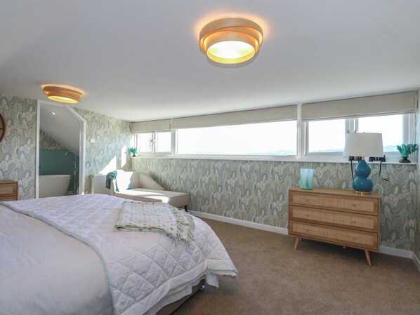A bedroom with a large bed, a dresser, a nightstand, a window with a view, and a bathroom with a bathtub and shower. - Swn Y Wylan (Pentraeth)