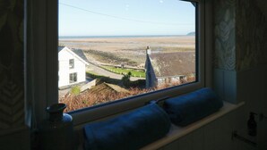 A room with a large window overlooking a beach and a white building. - Swn Y Wylan (Pentraeth)