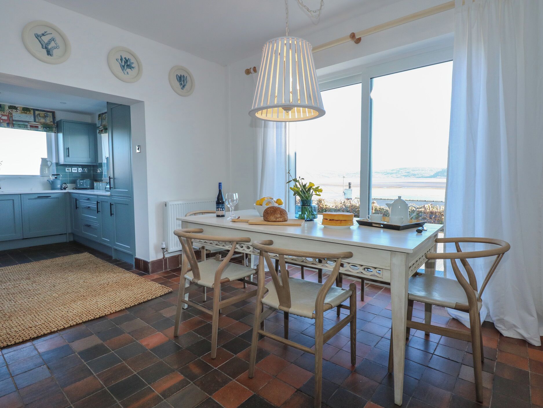 A kitchen with a dining area, featuring a large window with a view, a hanging light fixture, and a kitchen counter with various items.