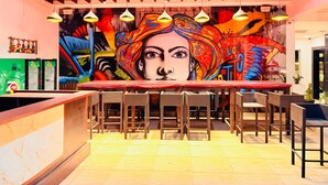 A vibrant bar area with a mural, wicker furniture, and a 7UP refrigerator. - Wow Hotels (Tirupur)