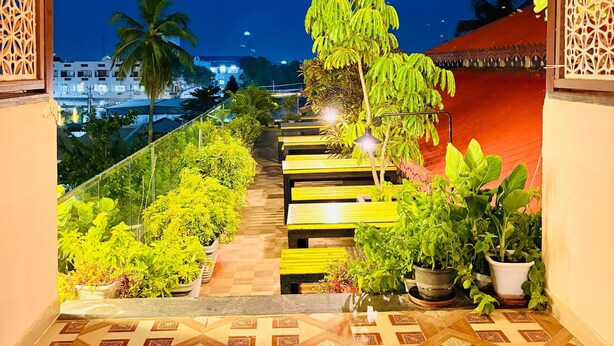 A nighttime view of a garden pathway with benches and potted plants, leading to a building with a red roof. - Wow Hotels (Tirupur)