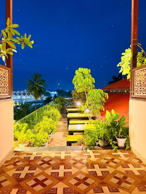 A nighttime view of a garden pathway with benches and potted plants, leading to a building with a red roof. - Wow Hotels (Tirupur)