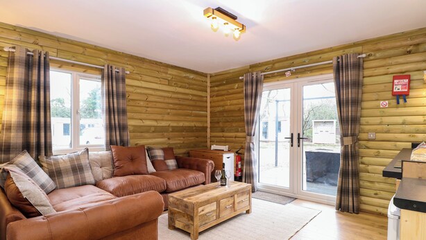 A cozy room with a brown leather sofa, a wooden coffee table, and plaid curtains. - Rabbit Warren Lodge (King's Lynn)