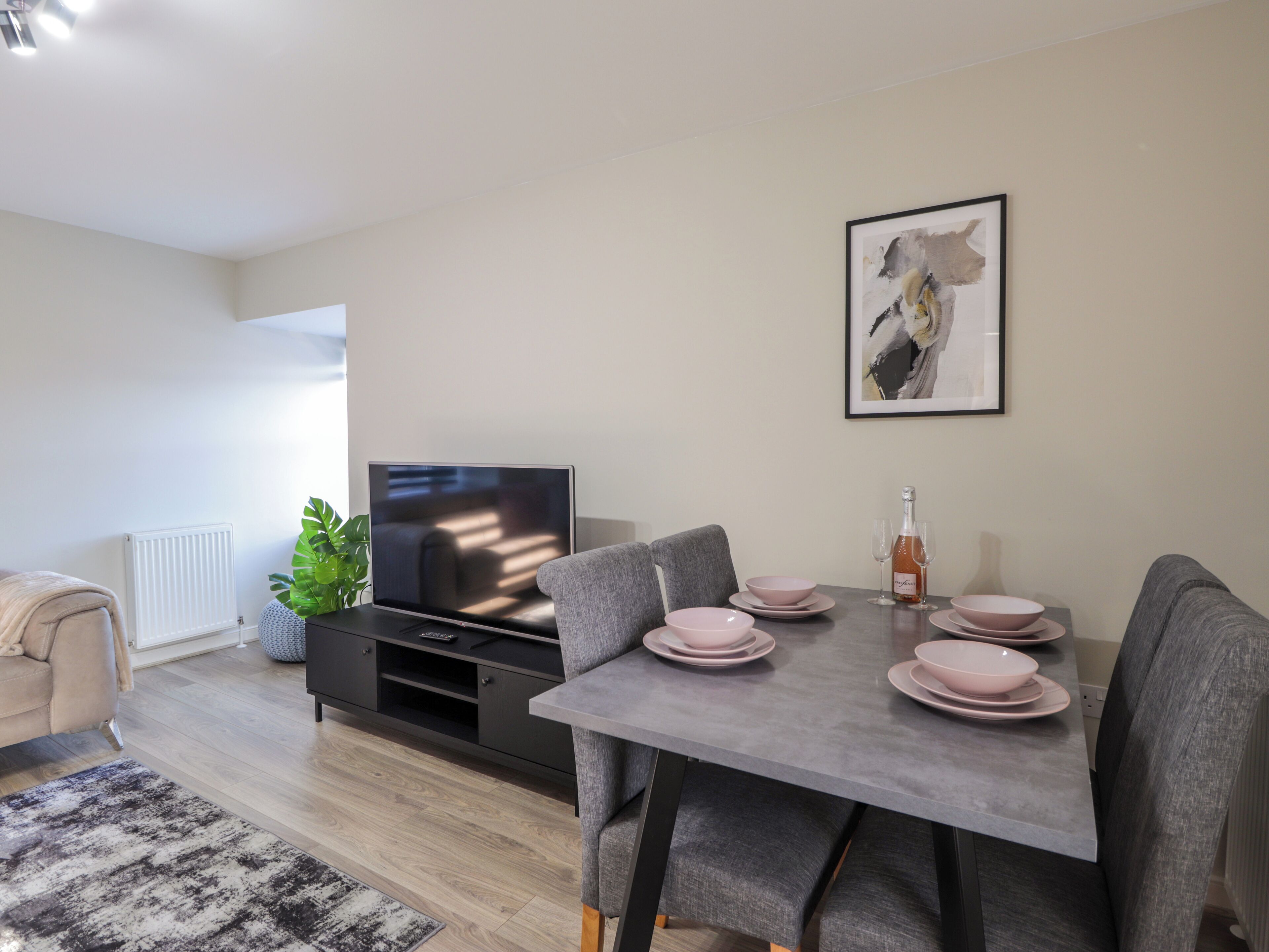 Una sala de estar moderna con una mesa de comedor para cuatro, un televisor de pantalla plana, un sofá gris y una obra de arte enmarcada en la pared.