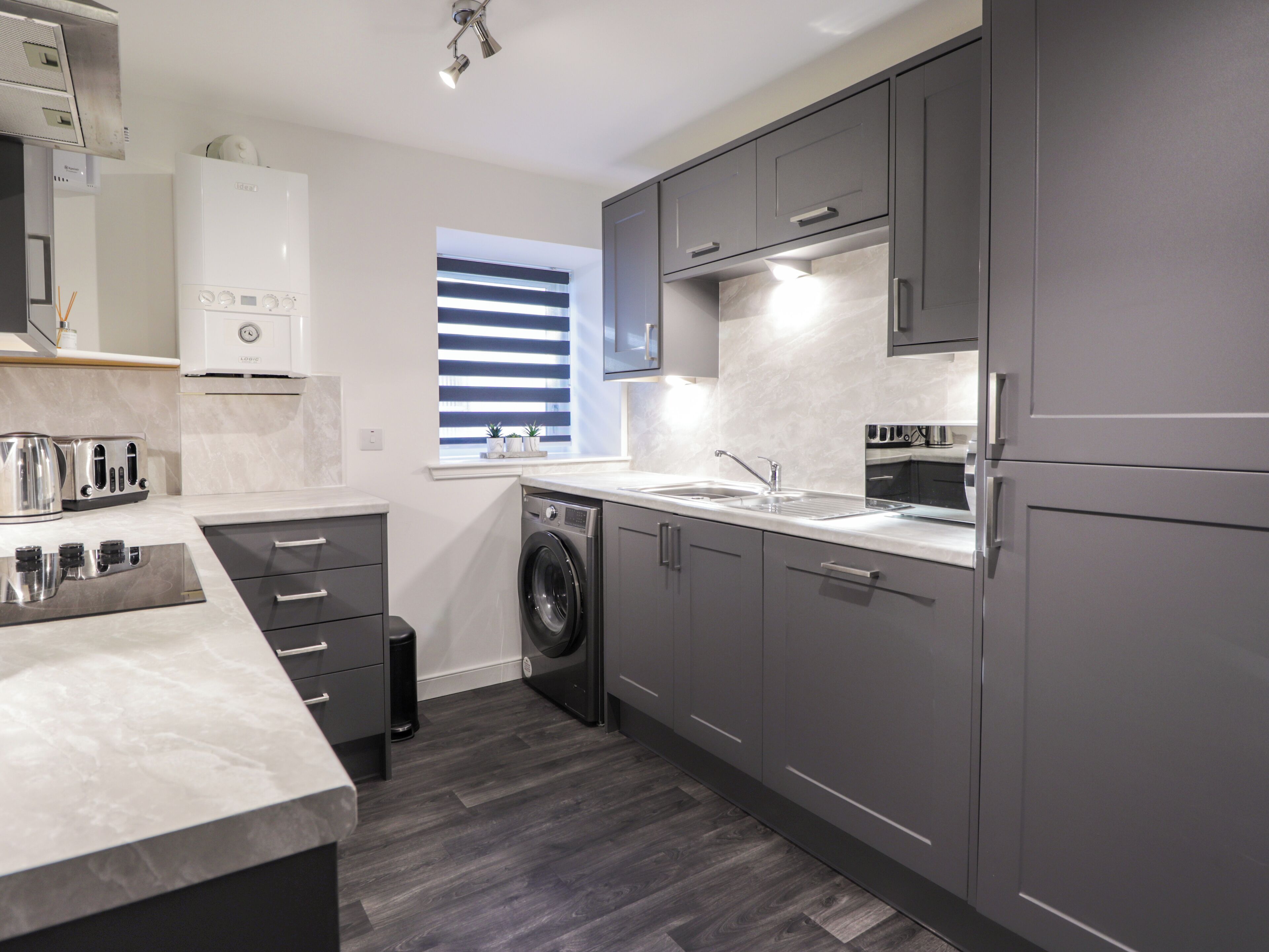 A modern kitchen with grey cabinets, a built-in oven, a washing machine, and a window with blinds.