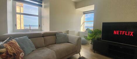 A living room with a sofa, a TV, and a view of the ocean.