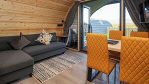 A cozy cabin interior with a wooden ceiling, a sofa with cushions, a dining table with chairs, and a large window offering a view of the outdoors. - Barn Owl (Carnforth)