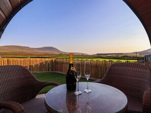 A table with a bottle of wine and two glasses, set against a scenic view of rolling hills and a wooden fence.