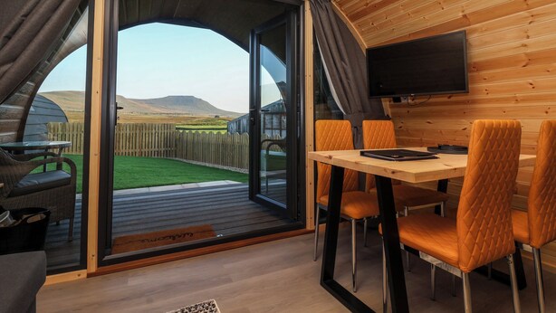A cozy interior with a wooden ceiling, a dining area with orange chairs, and a large sliding glass door leading to an outdoor space with a wooden fence and a grassy lawn. - Barn Owl (Carnforth)