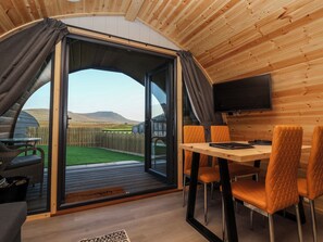 Un interior acogedor con techo de madera, una zona de comedor con sillas anaranjadas y un amplio ventanal corredera que da a un espacio exterior con valla de madera y césped.