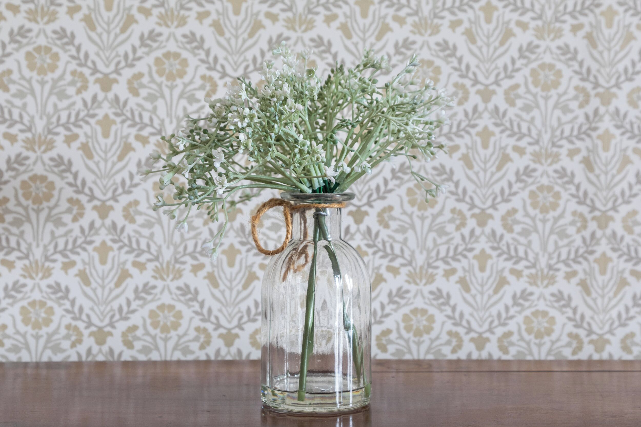 Un florero de vidrio transparente con asa, lleno de follaje verde, sobre una superficie de madera frente a un fondo de papel tapiz estampado.
