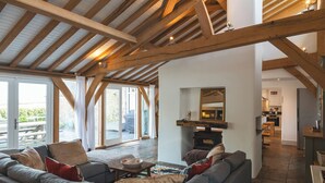 A spacious living area with a large gray sofa, wooden beams, a fireplace, and a view of the outdoors through large sliding glass doors. - The Farmhouse at Broad Downs (Kingsbridge)