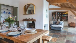A dining area with a wooden table set for a meal, a fireplace, and a cozy living room with a sofa and bookshelves. - The Farmhouse at Broad Downs (Kingsbridge)